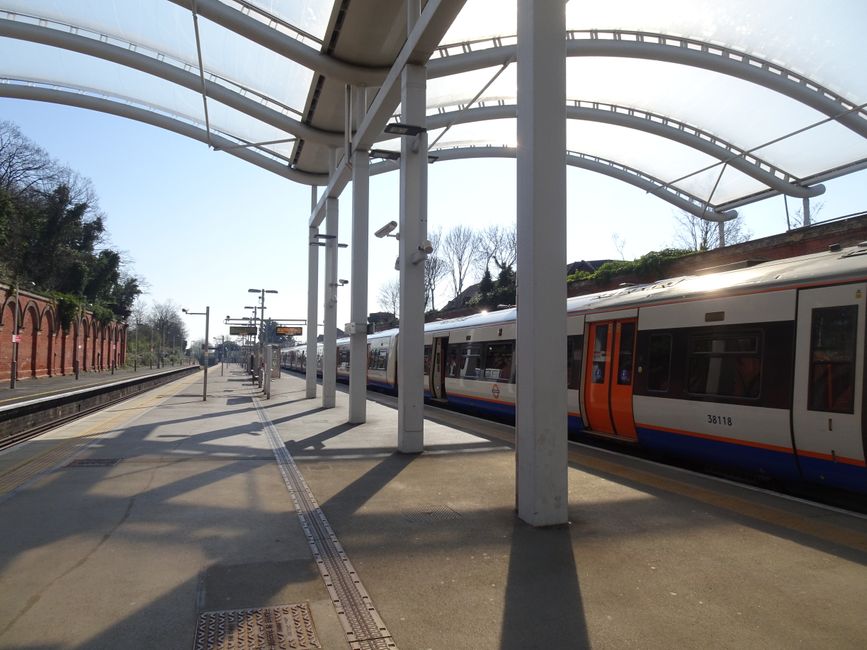 The image shows an outdoor train station platform with a modern train arriving. The platform surface is made of concrete and features tactile paving strips along the edge for safety. Overhead, a metal